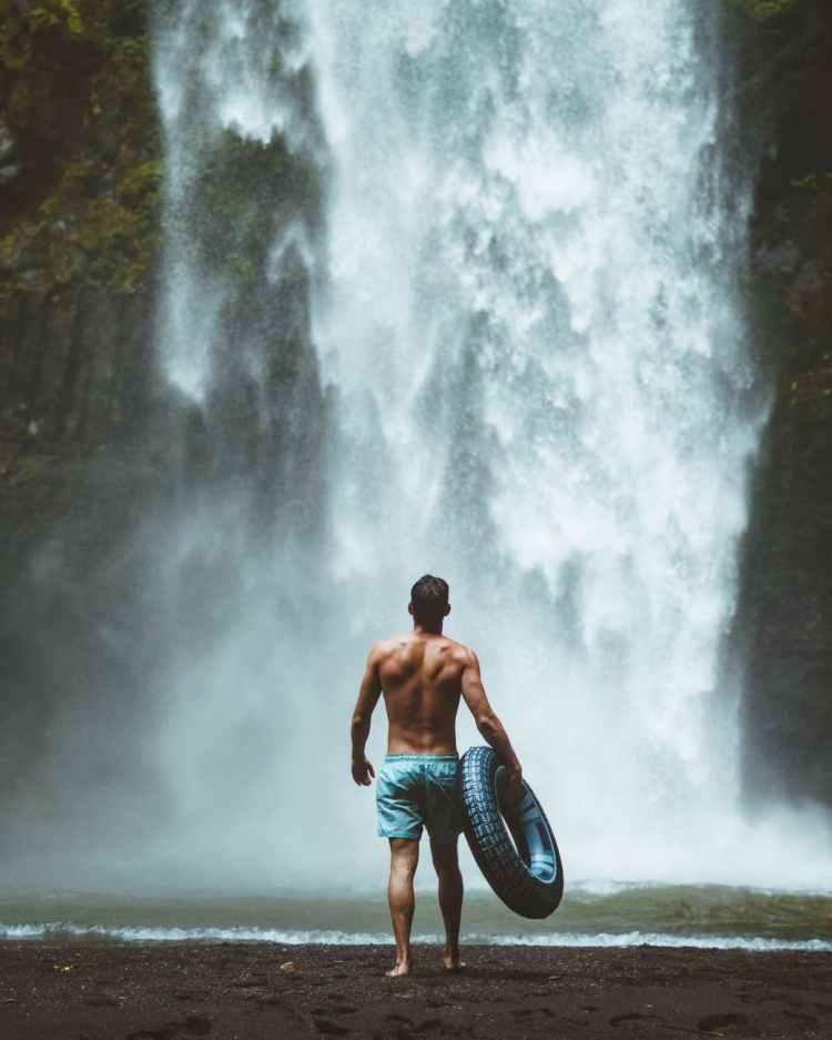 man wearing blue shorts holding vehicle tire facing waterfalls
